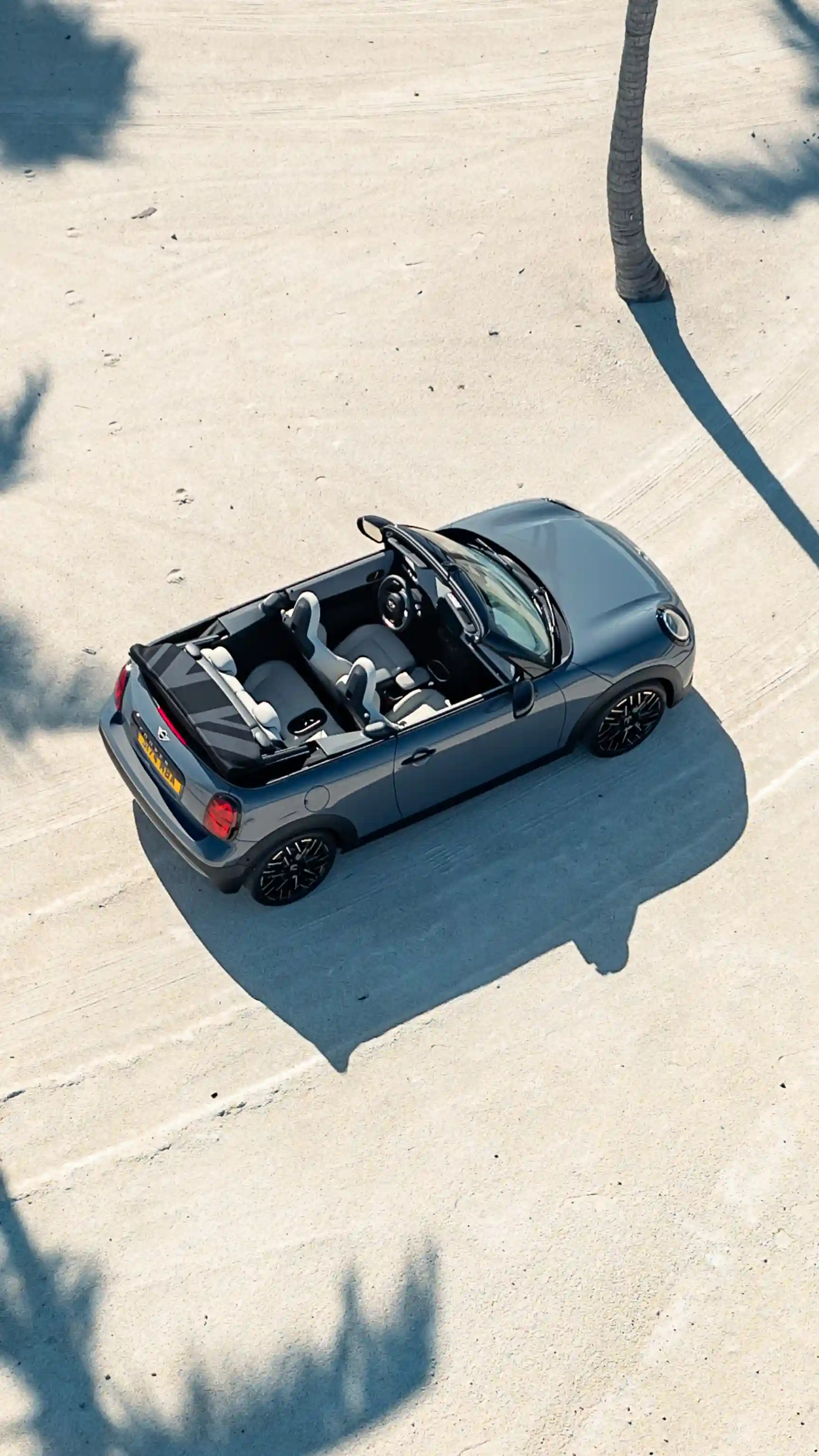 Person sitting on the open boot edge of a MINI Convertible with luggage packed in the rear, while another person walks nearby.