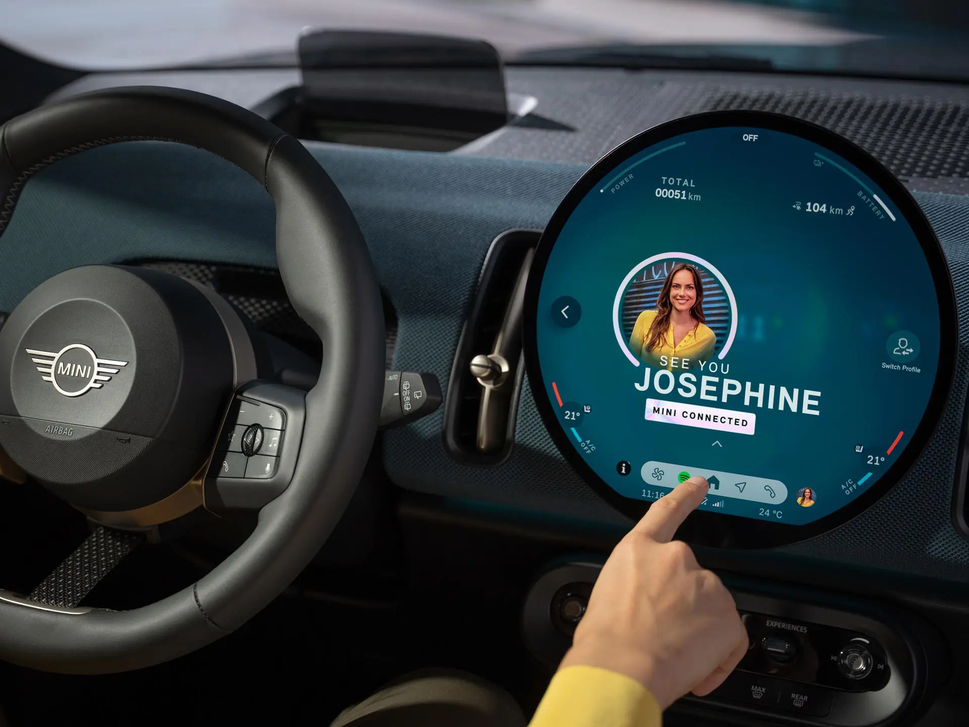 Driver interacting with the circular MINI infotainment display showing a MINI Connected profile screen.